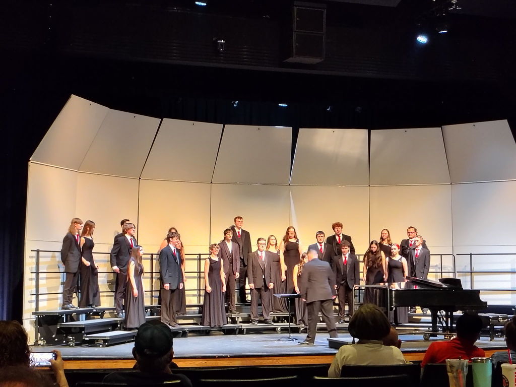 A group of performers in formal attire stand on a stage with musical instruments in front of an audience.