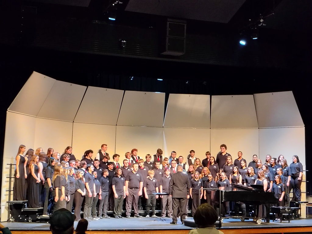 A large choir dressed in matching shirts stands on a stage, facing an audience, with a piano at the center.