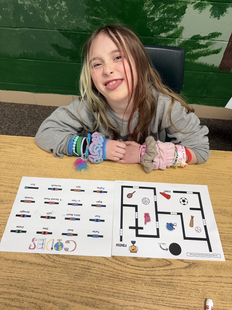 Child seated at a table with a paper maze, smiling, and leaning on the table with their elbows.