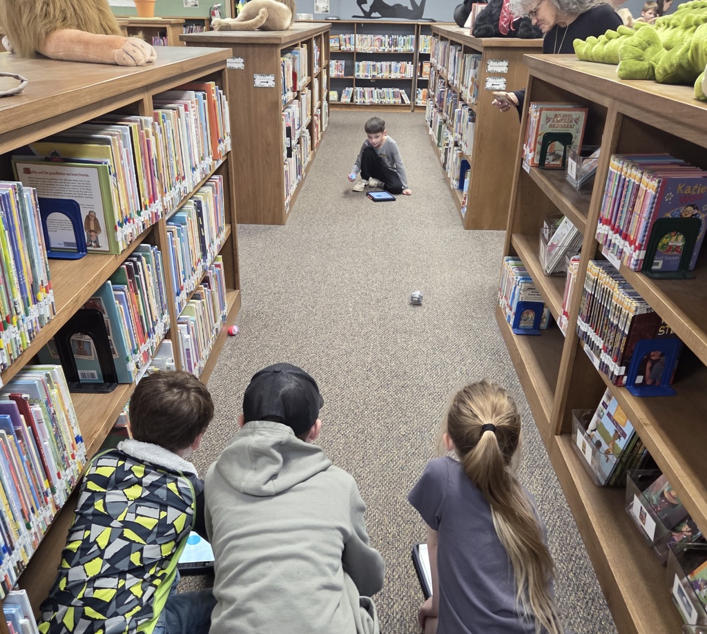 Library aisles with wooden shelves filled with books. Children sit on the floor reading tablets. A boy sits in the aisle using a tablet.