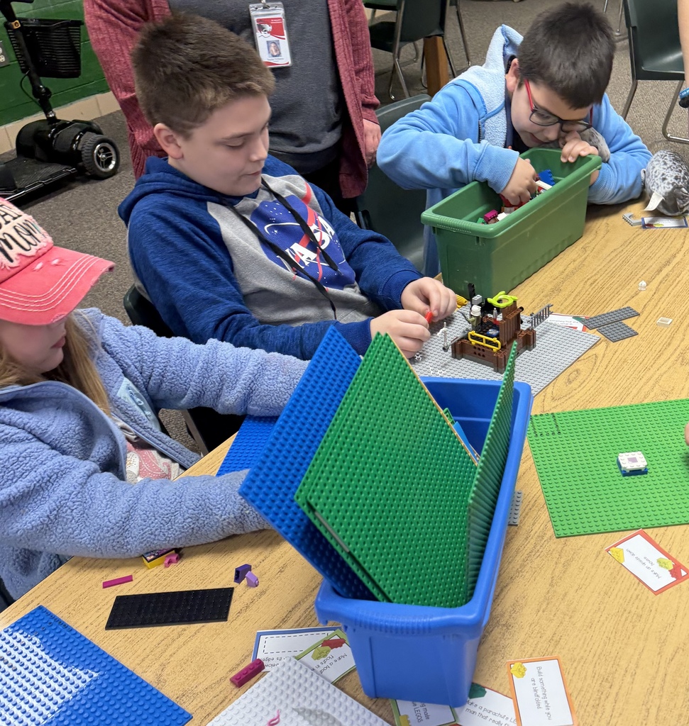 Four children, three boys and one girl, work on Legos at a table. One child wears a pink hat.