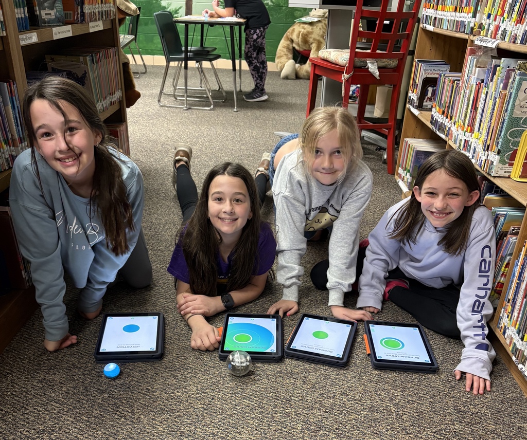 Four girls in sweatshirts sit in a library with tablets on the floor, smiling at the camera.