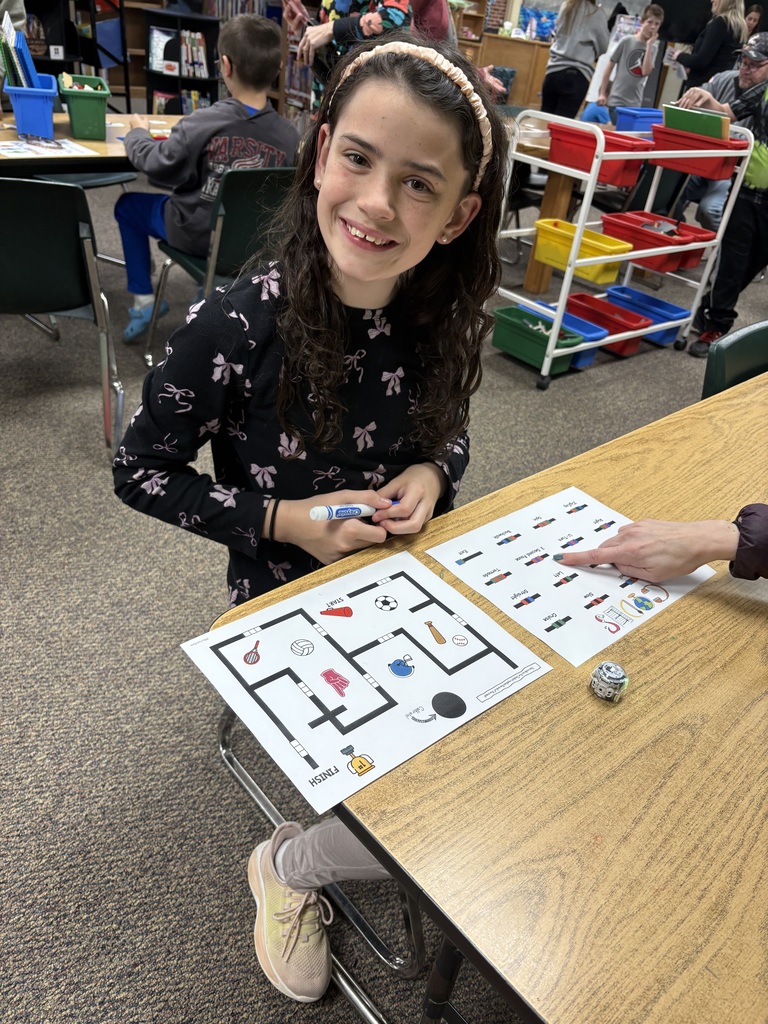 A child sits at a table, smiling, and holds a marker. In front is a maze puzzle with a small robot.