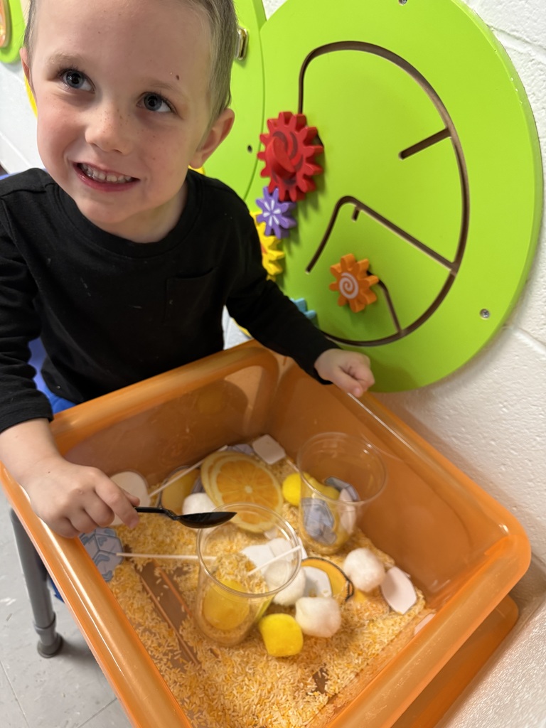 A child holds a spoon in a sensory bin filled with dry ingredients and fruit.