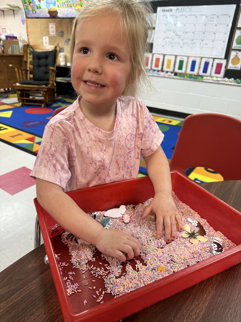 A young girl sits in a chair at a table. She is smiling while touching multicolored items in a tray.