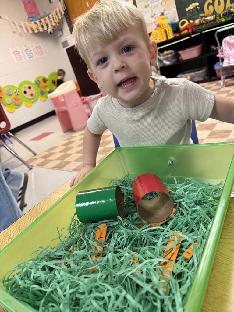 A young child in a white t-shirt reaches into a tray filled with green grass and red and green objects.