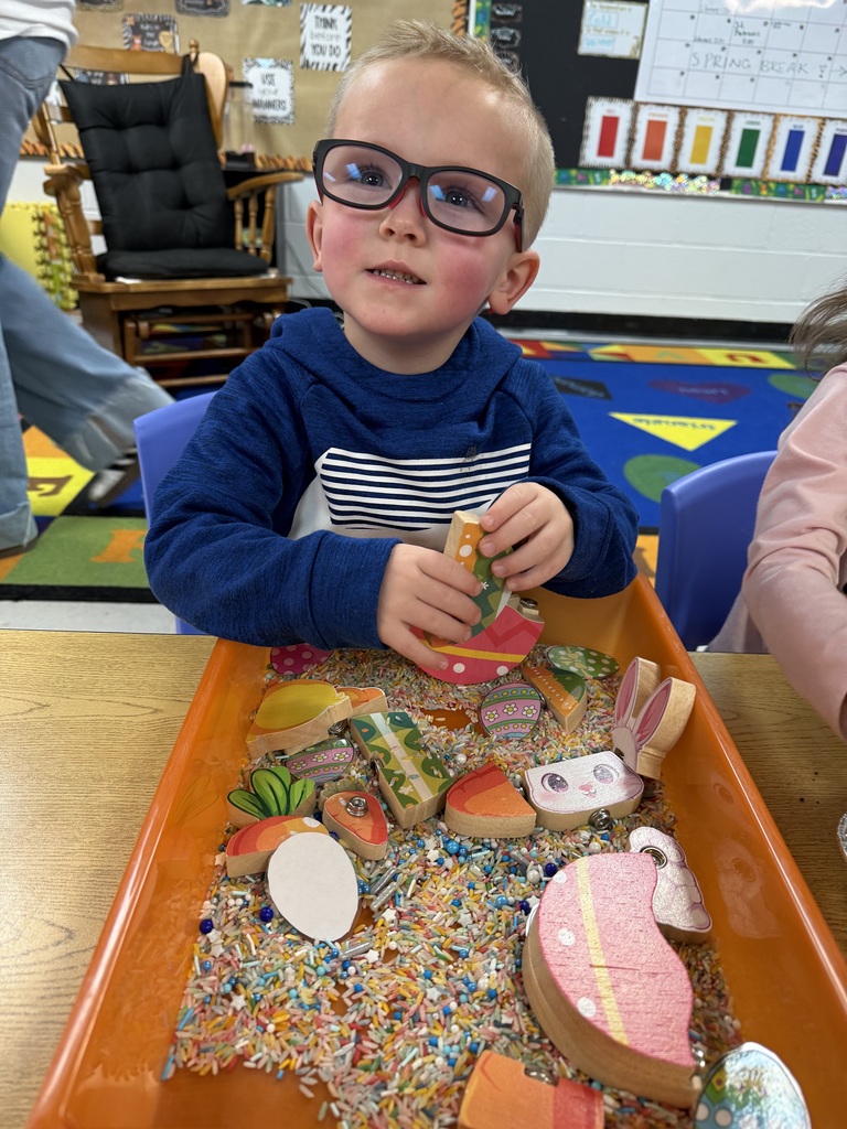 A young child in glasses sits in a chair and handles objects in a tray.