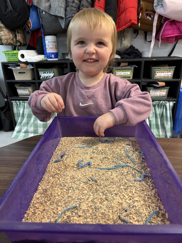 Child in a purple tray filled with sand and blue yarn in a classroom.