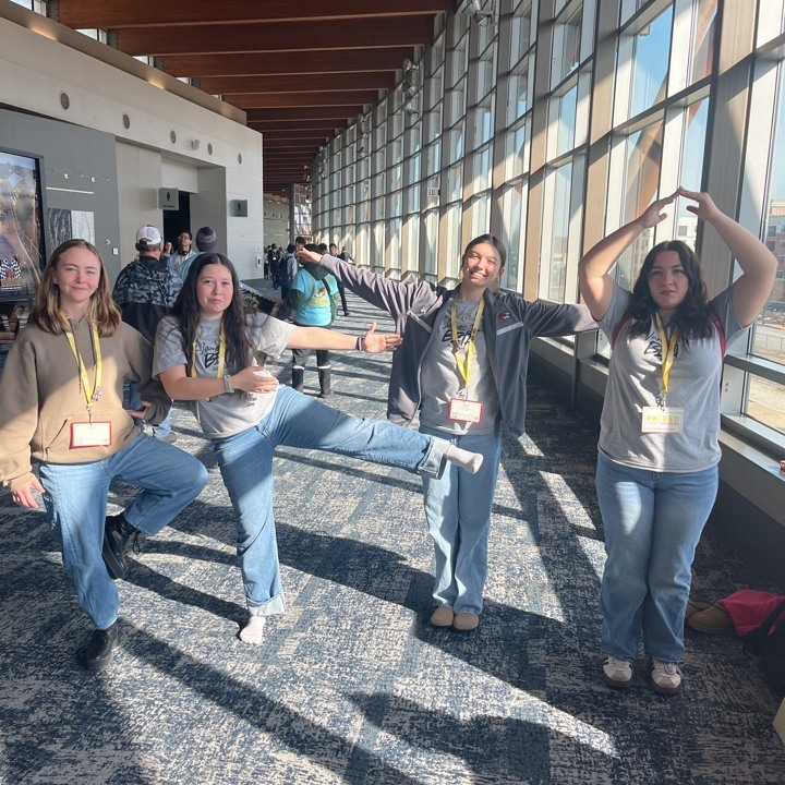 Four women in jeans and ID lanyards pose for a photo in a hall with large windows.