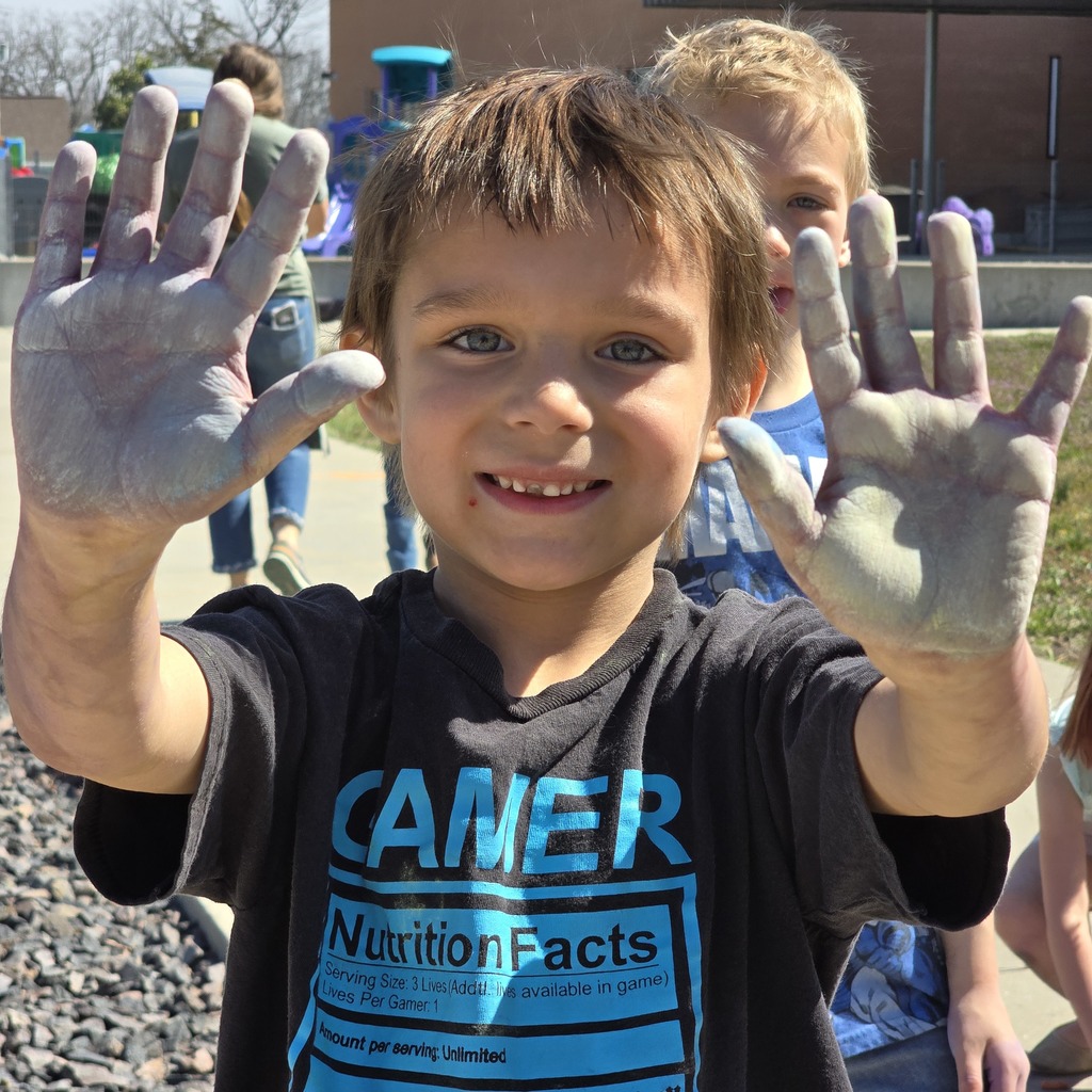 A boy with dirt on his hands smiles and shows his palms. He wears a dark shirt with "CAMER" and "Nutrition Facts" printed on it.