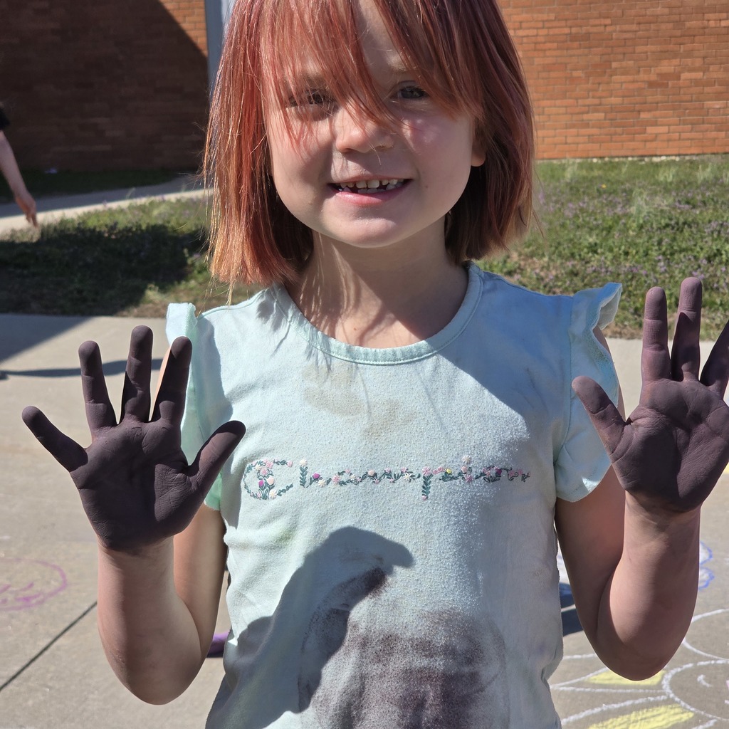 Young girl with red hair, blue shirt, and brown paint on her hands stands on sidewalk.