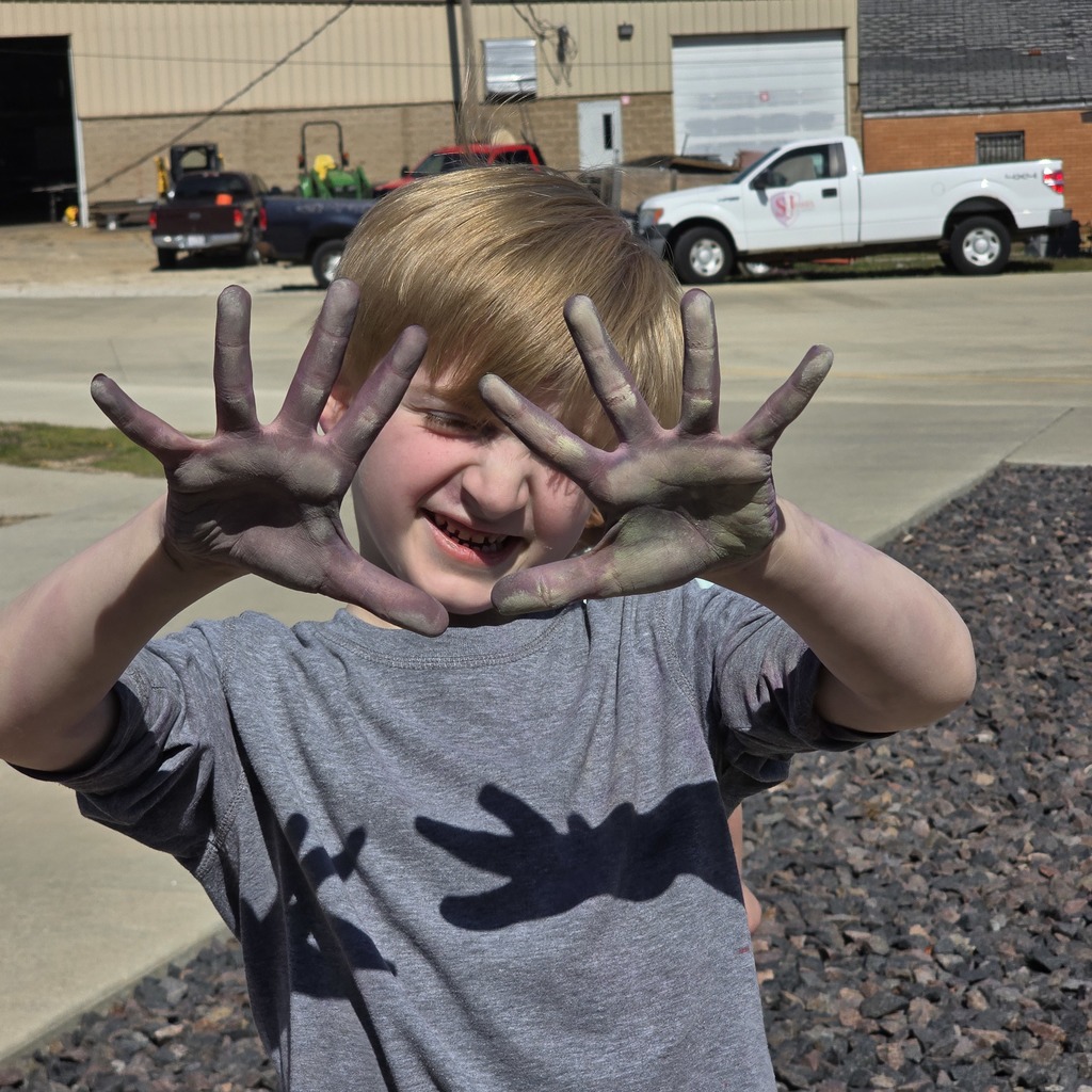 A child with hands covered in green paint smiles at the camera. Behind the child are vehicles and a building.