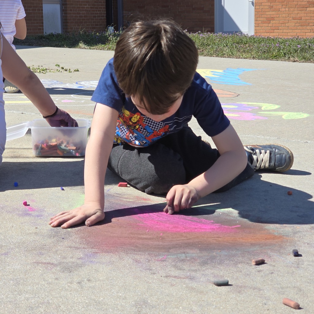 A child crouches on concrete, drawing with chalk. A hand, partially visible, holds a container of chalk nearby.