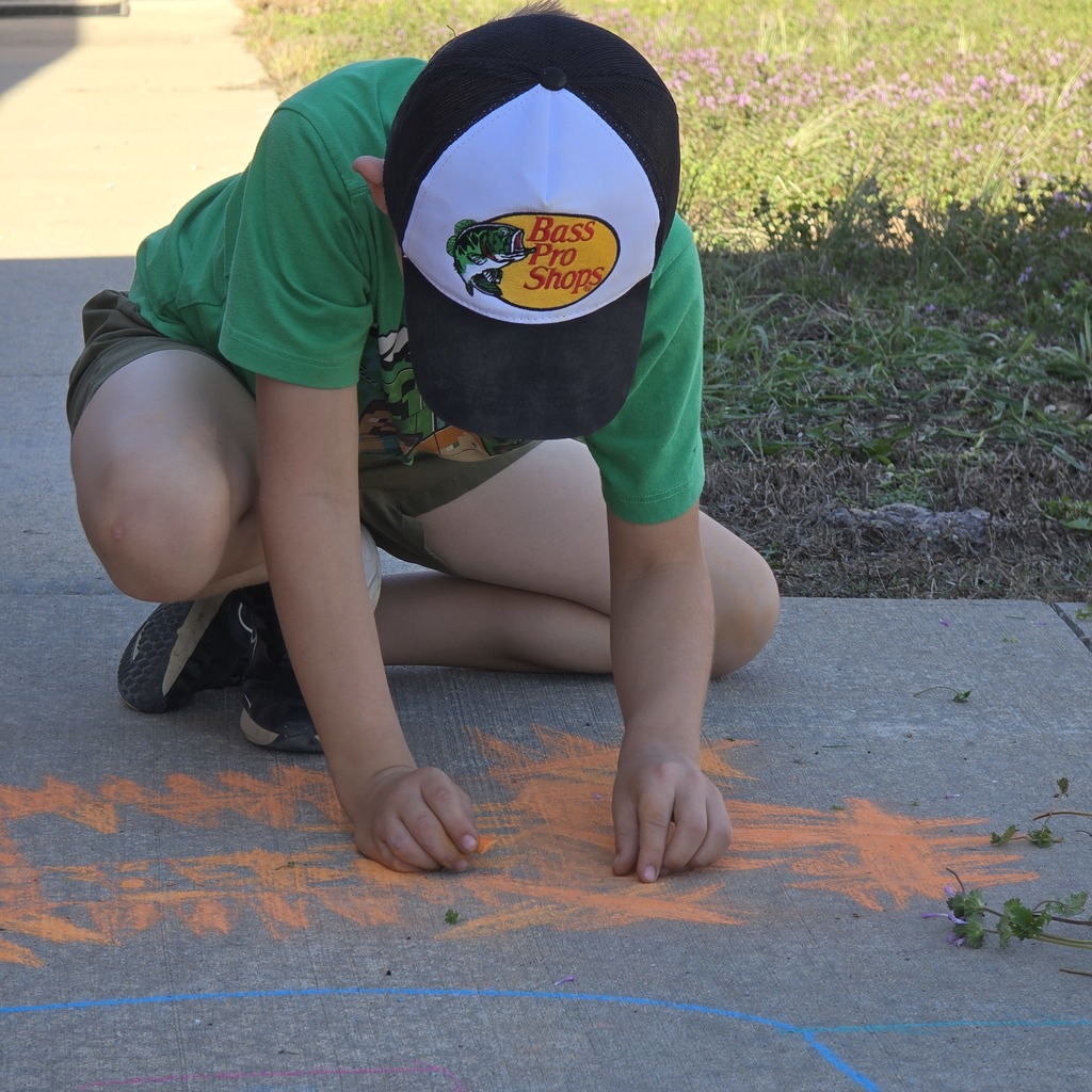 A person in green and black cap draws a fish shape on the sidewalk with orange chalk.