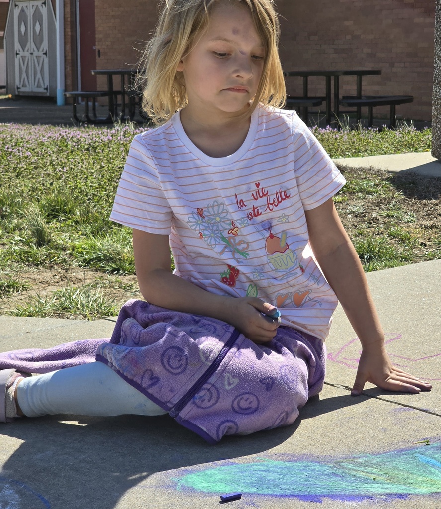 A young girl with blonde hair sitting on a sidewalk, drawing with chalk. Behind her is a building with brick walls.