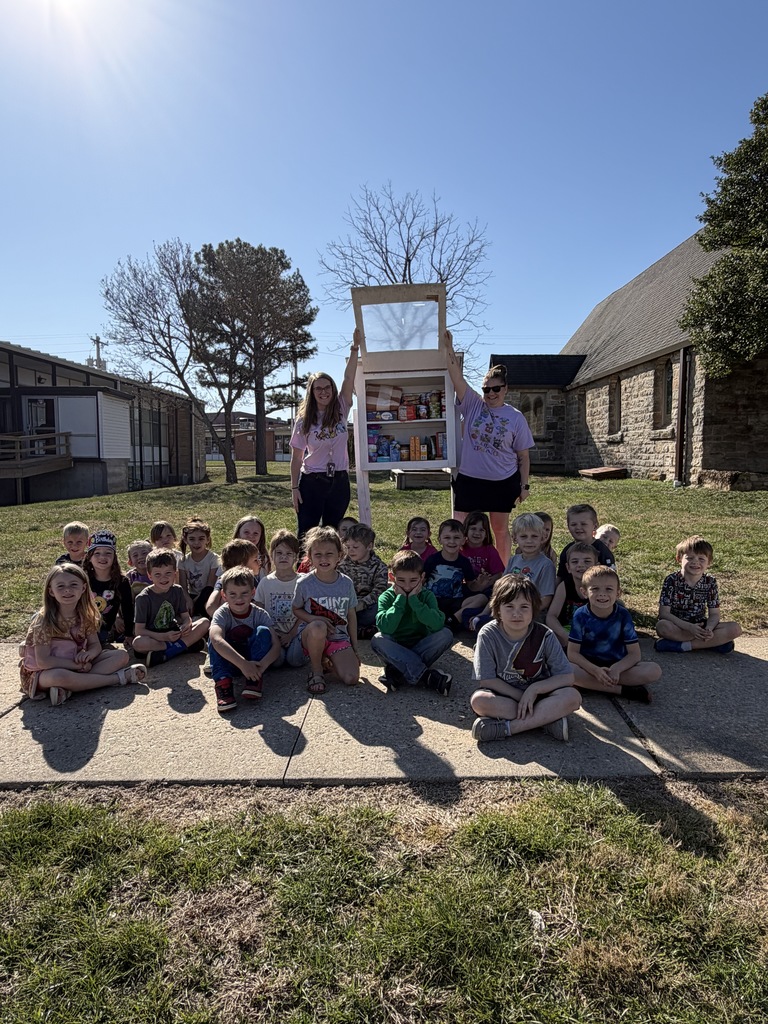 Group of children sitting on sidewalk, with two adults holding a box. Behind them, a building and trees.