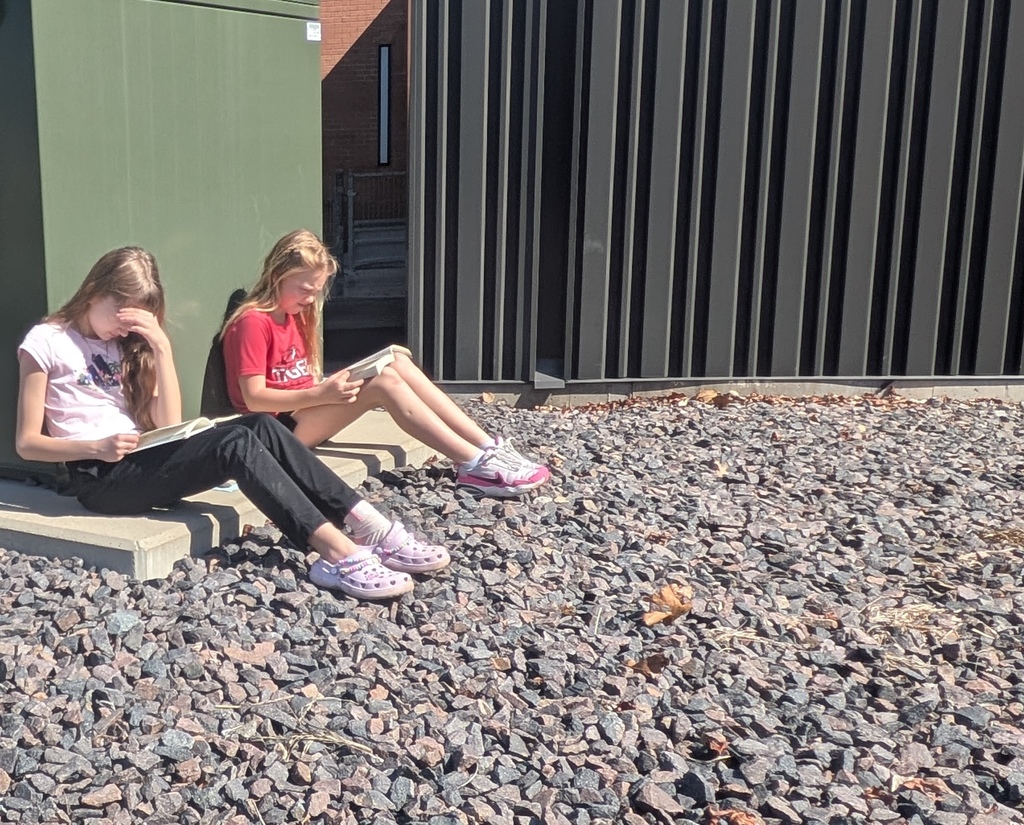 Two young girls sit on concrete steps, holding books, with gravel ground and a green building behind them.