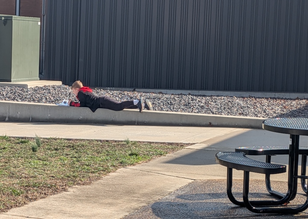 Person lying on concrete barrier with red top, near metal wall, gravel, and picnic table.