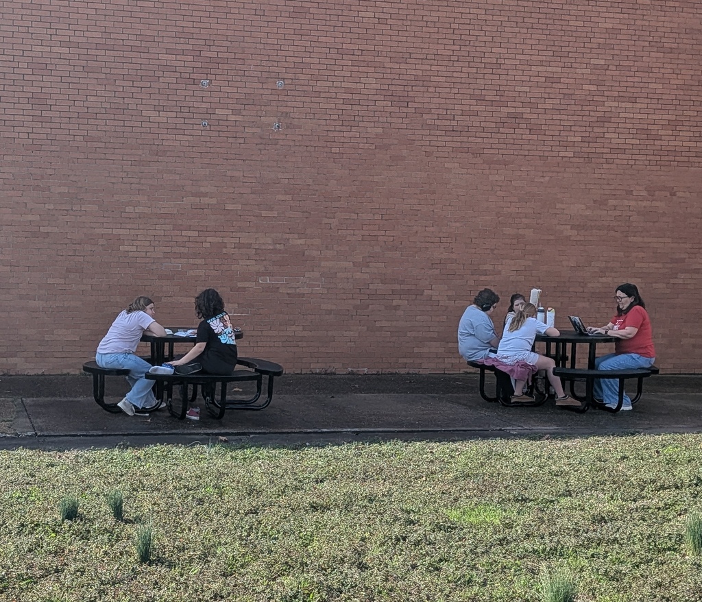 Four people seated at tables, one using a laptop, on a concrete walkway with brick walls.