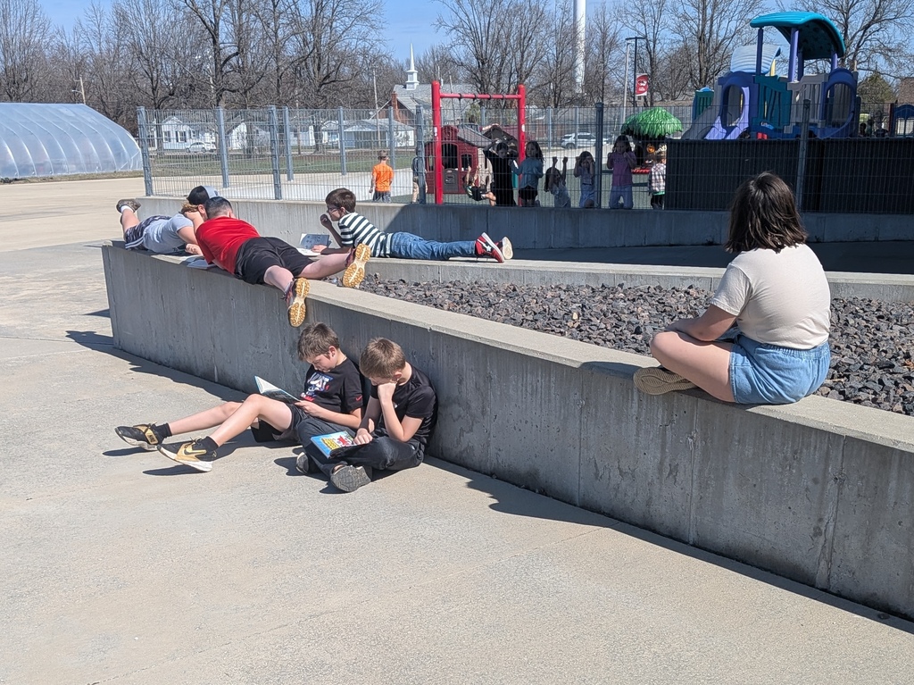 Several people sit on a concrete wall near a playground. A group of children sits on the ground.