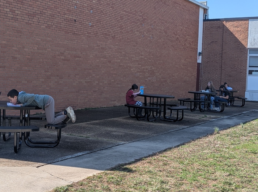 Students sit at picnic tables outside a brick building with a concrete pathway, some reading or resting.