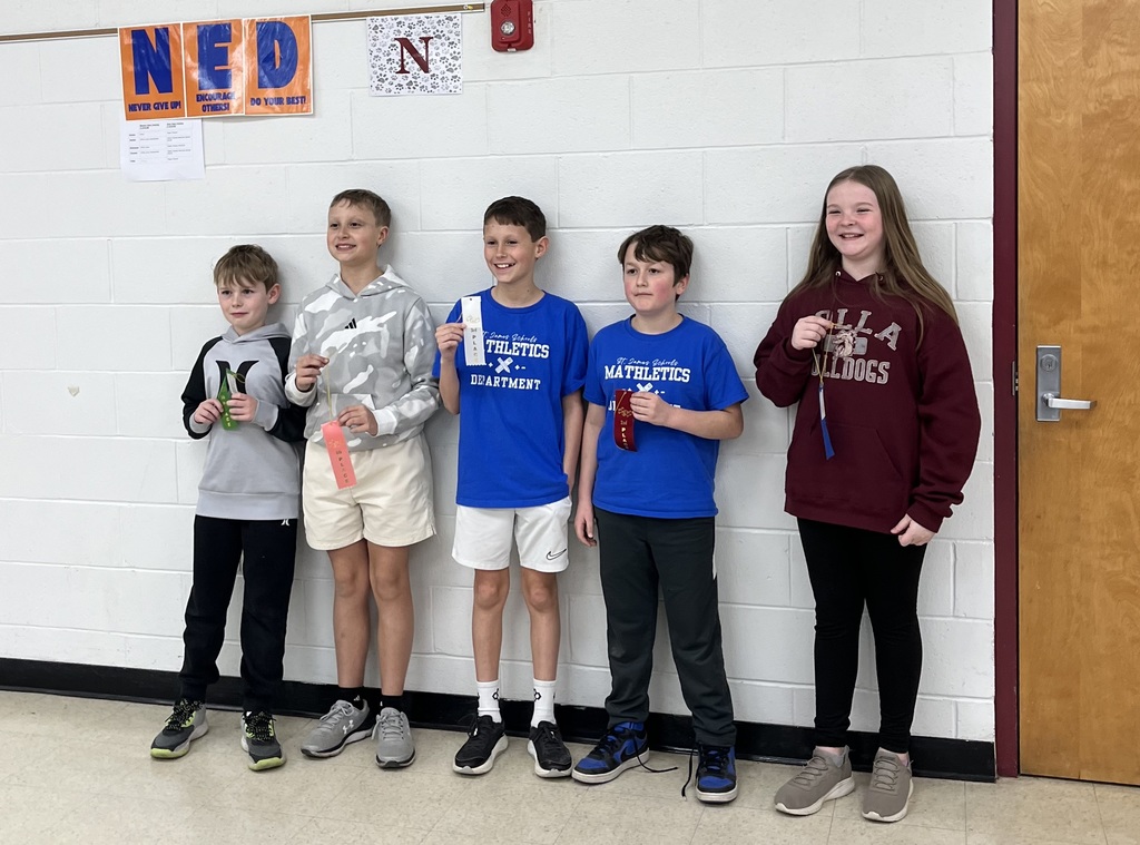Five children stand together, holding awards, with one in a silver shirt, three in blue, and one in maroon. They are positioned in front of a white wall with orange signs.