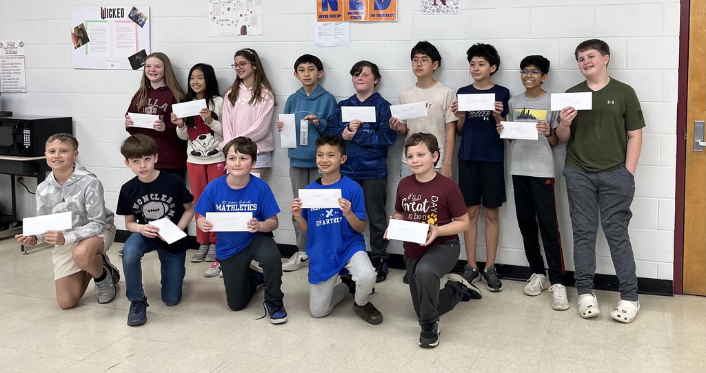 A group of children hold certificates and pose for a photo in a classroom with a white wall.