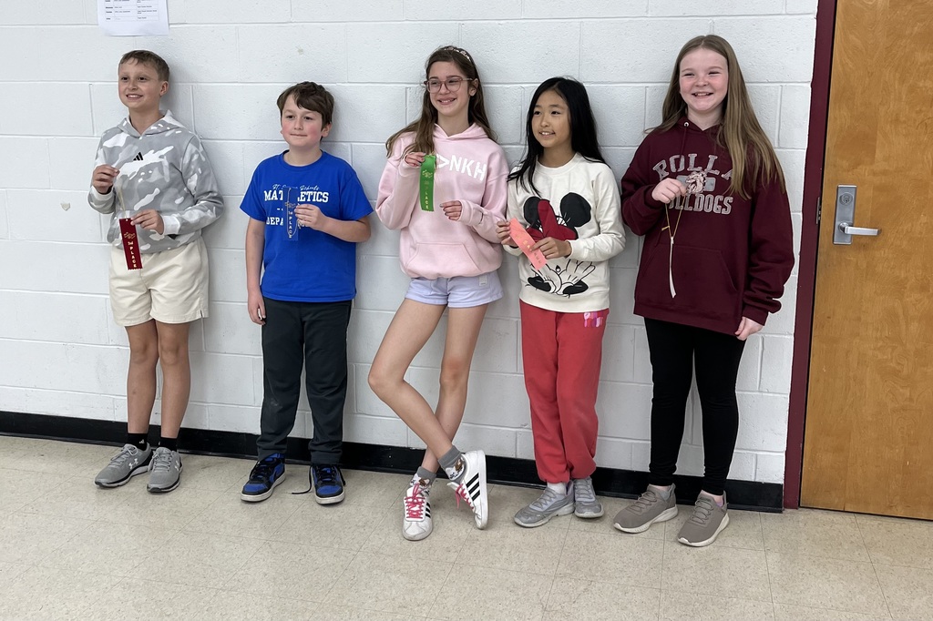 Five children stand in a row near a door, holding awards. They wear casual clothes. One wears a pink jacket with a cartoon character.