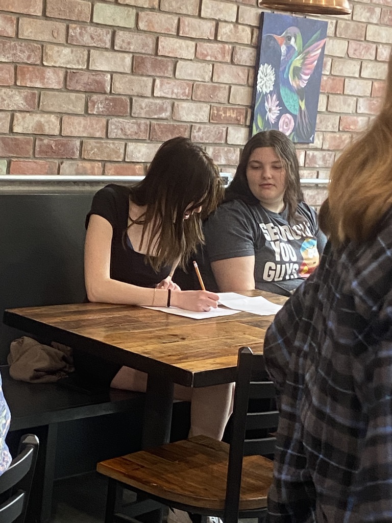Two women seated at a wooden table. One writes on paper while the other observes. A brick wall with artwork in the background.