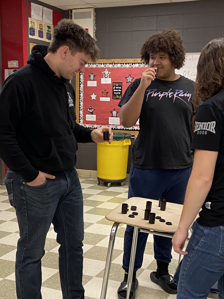 Three young people in black tops and jeans play chess on a table in a classroom.