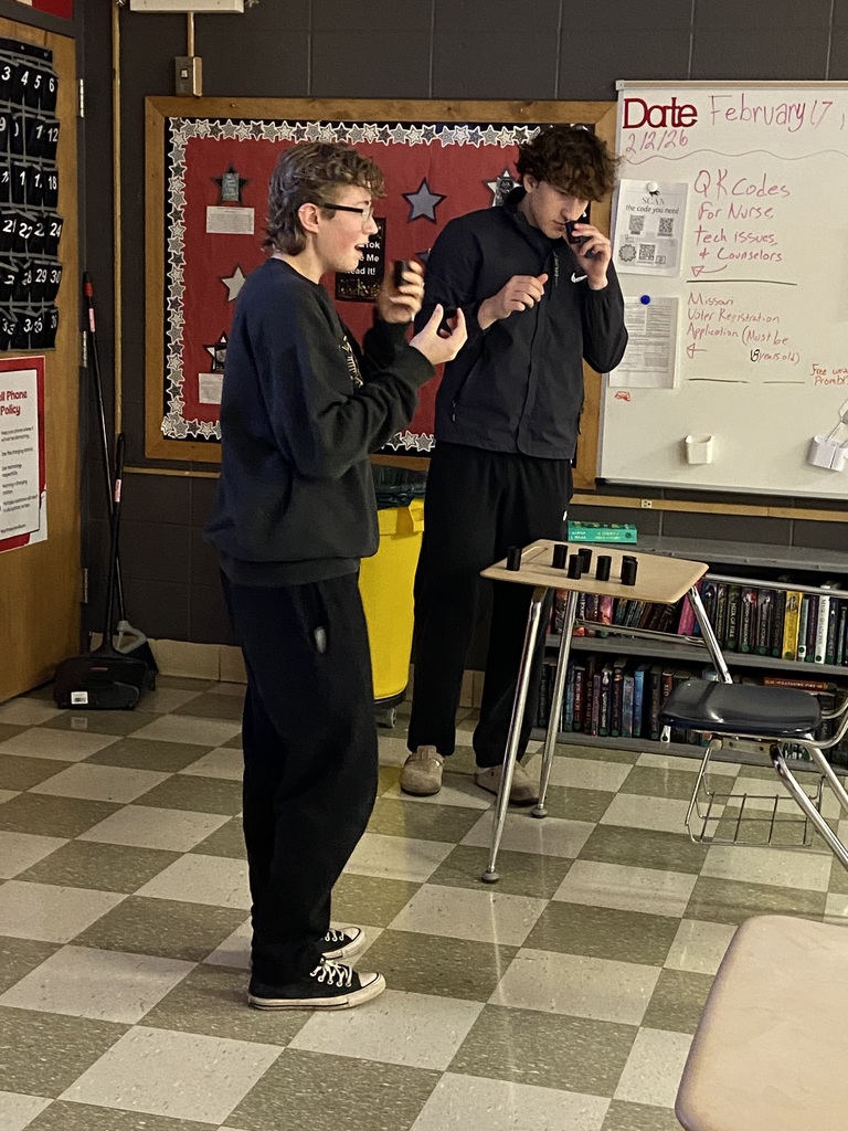 Two individuals in a classroom are playing a board game. One is in a black shirt, the other in a blue jacket.