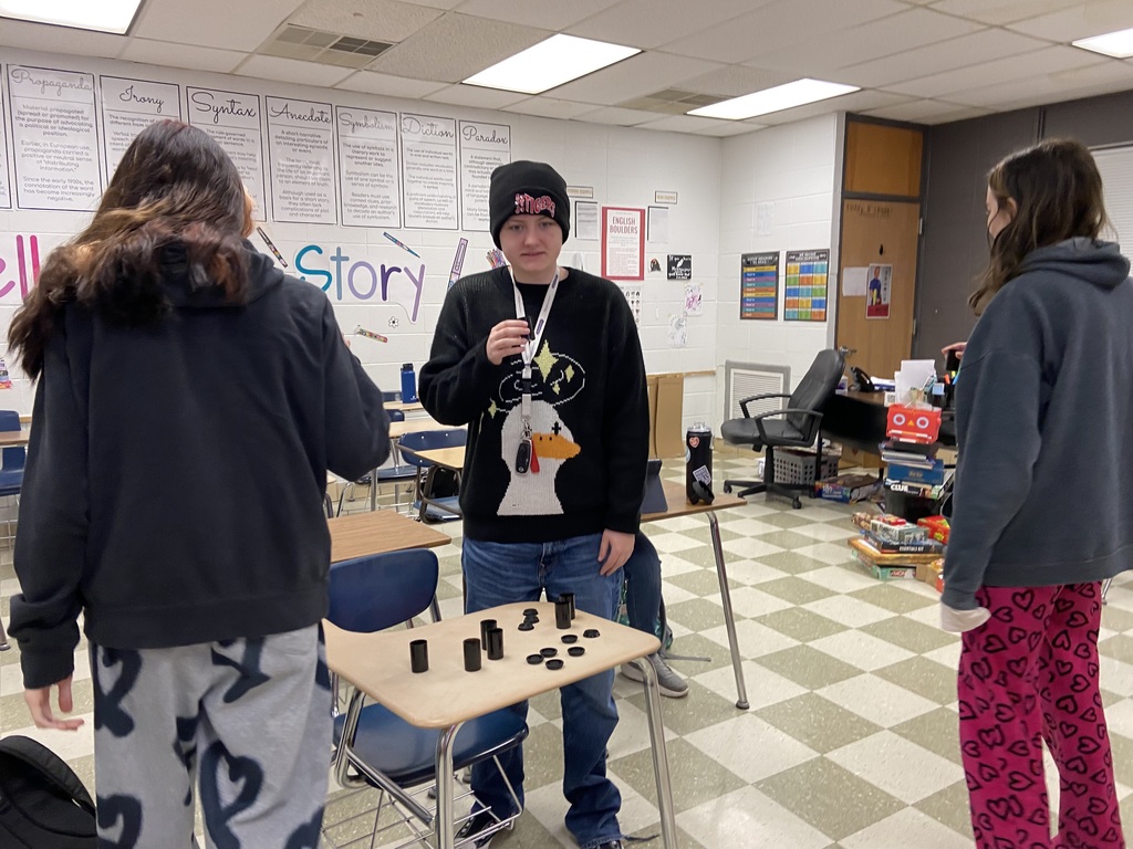 A person stands between two others, holding a chess set. They are in a classroom with checkered floors.