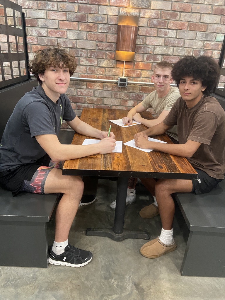 Three people sit at a wooden table, each with a pen and paper, against a brick wall.