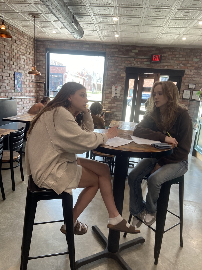 Two women sit at a small table in a coffee shop. One holds a pen and paper.