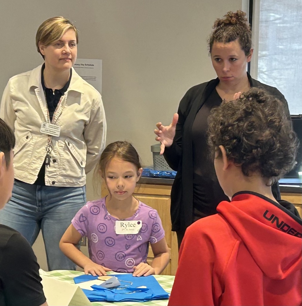 People in a room, one girl sits on a table, others stand. The woman on the right wears a black blazer, the woman on the left wears a white jacket.