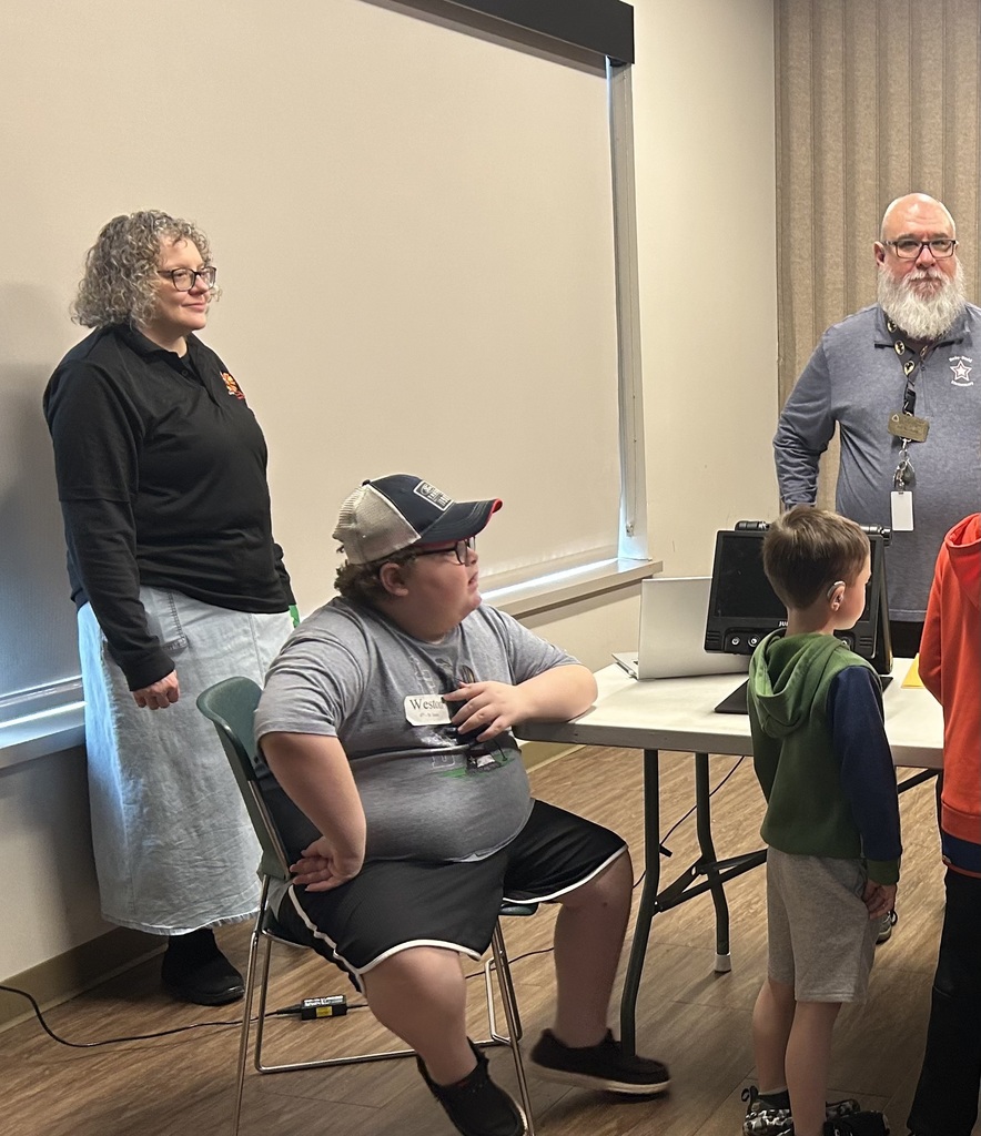 A group of people gathered in a room with a white wall, including a seated man with glasses and a woman standing.