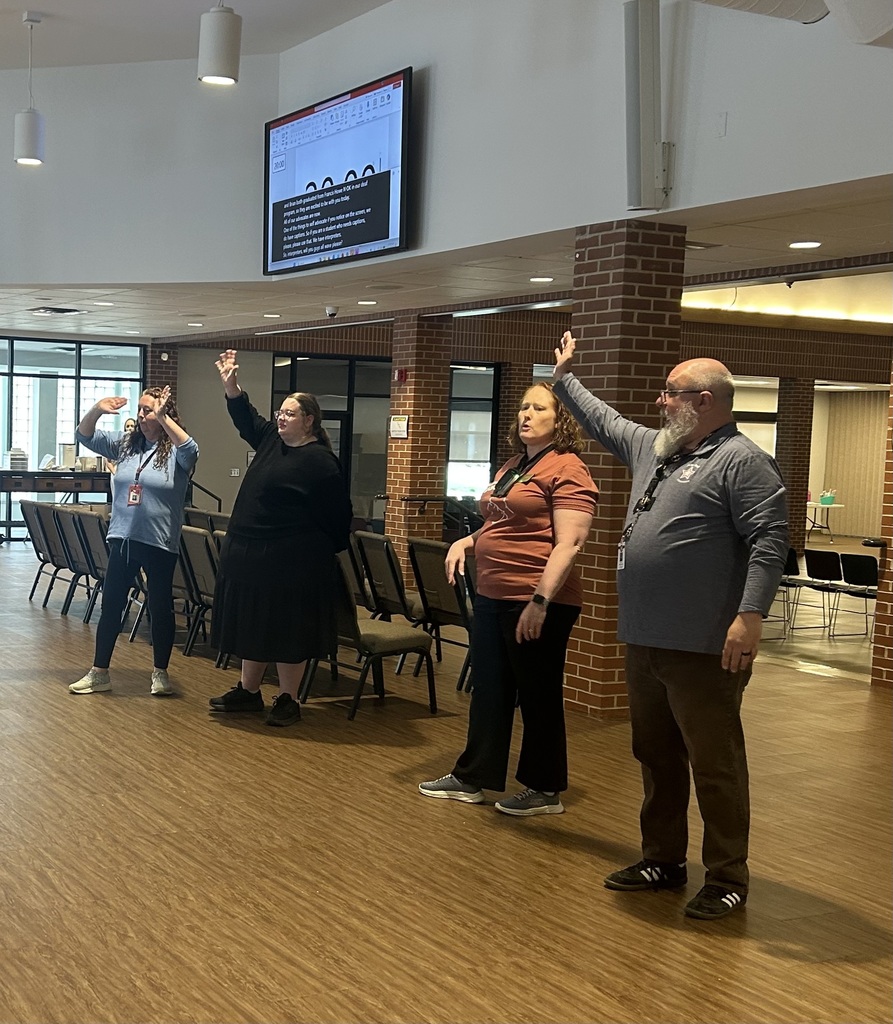 Four adults stand in a room with hands raised, facing a screen. Wooden floors and brick columns.