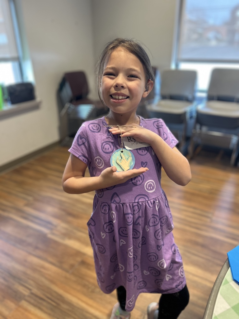 A young girl in a purple dress displays a circular medal. She stands in a room with wooden floors and chairs.