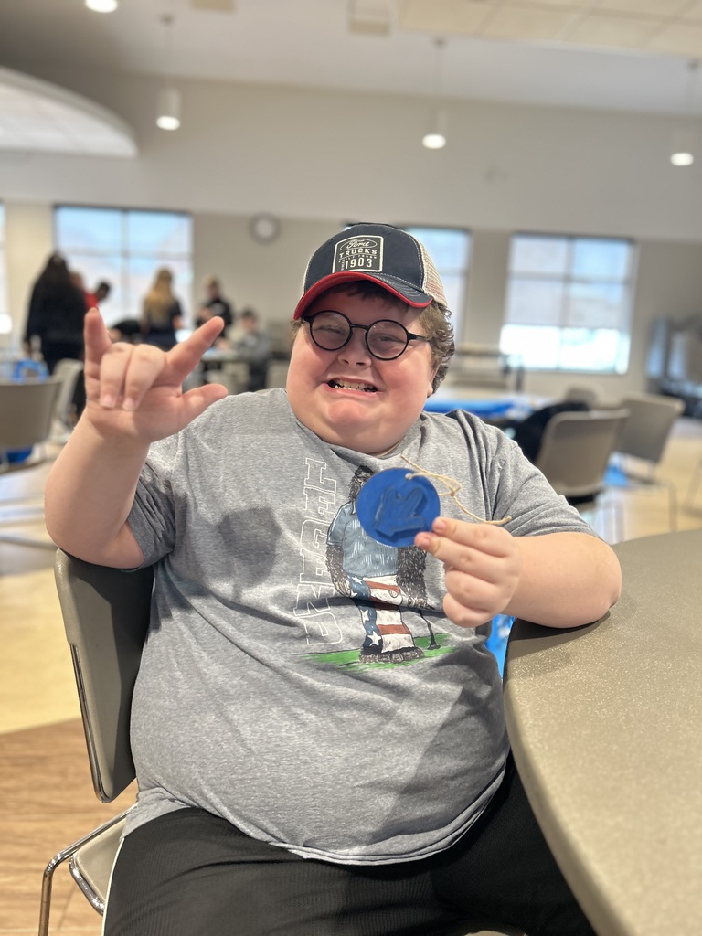 Person wearing a baseball cap, glasses, and a gray t-shirt sitting in a chair and holding a blue item.