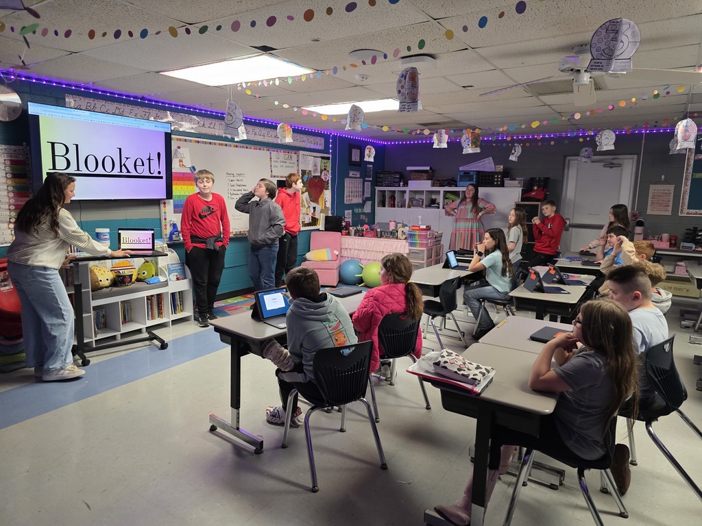 In a classroom, children are seated at tables with laptops. A teacher stands in front of a projector screen reading "Blooket!"