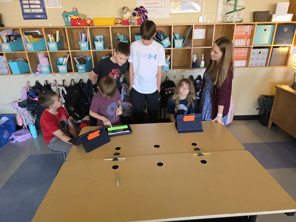 Children at a desk with holes and electronic devices, with shelves and storage in the background.