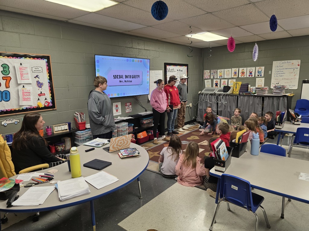 A classroom with tables, chairs, and a large screen. Some children are seated on the floor while others stand.