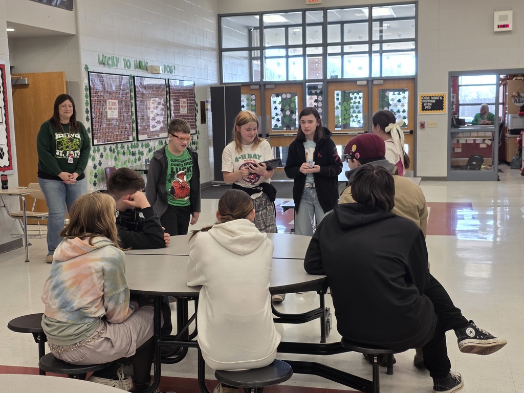 A group of people seated around a table, with some standing nearby, in a brightly lit cafeteria area with white walls and a glass door.