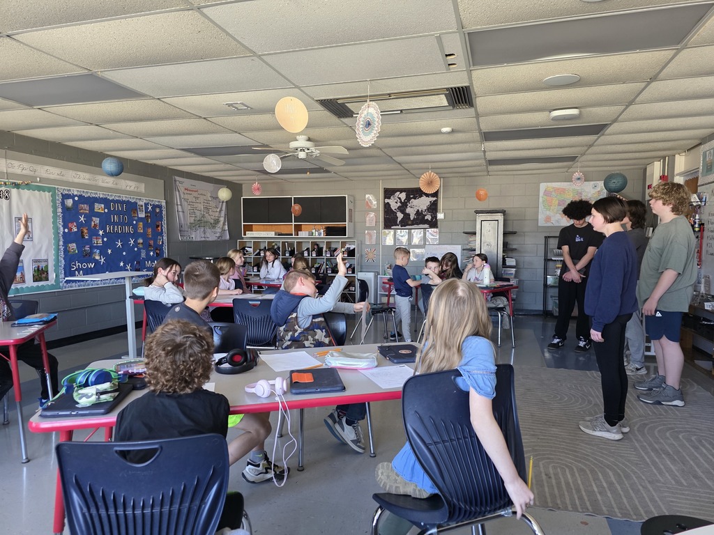 Classroom with students seated at desks. Some students are standing and others are raising their hands.