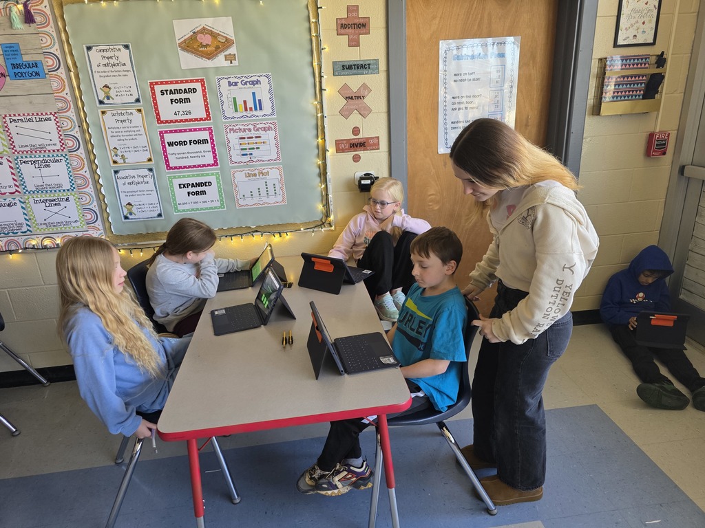 A teacher helps children at a table in a classroom with laptops. A bulletin board is in the background.