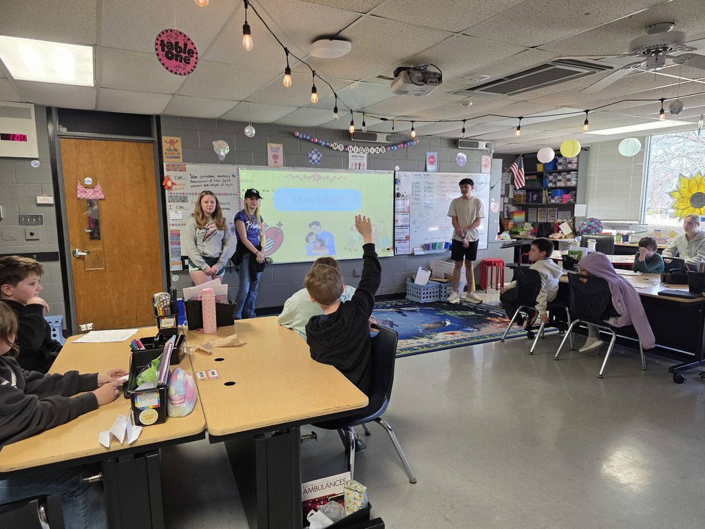 A classroom with students at tables and a projector screen. Some students are standing near the screen, others seated.