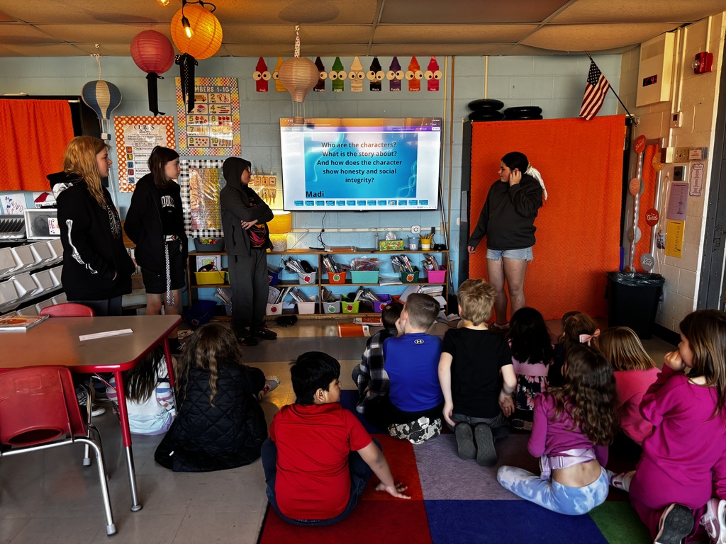 Several children sit on the floor in front of a teacher. A projector screen is visible in the background.