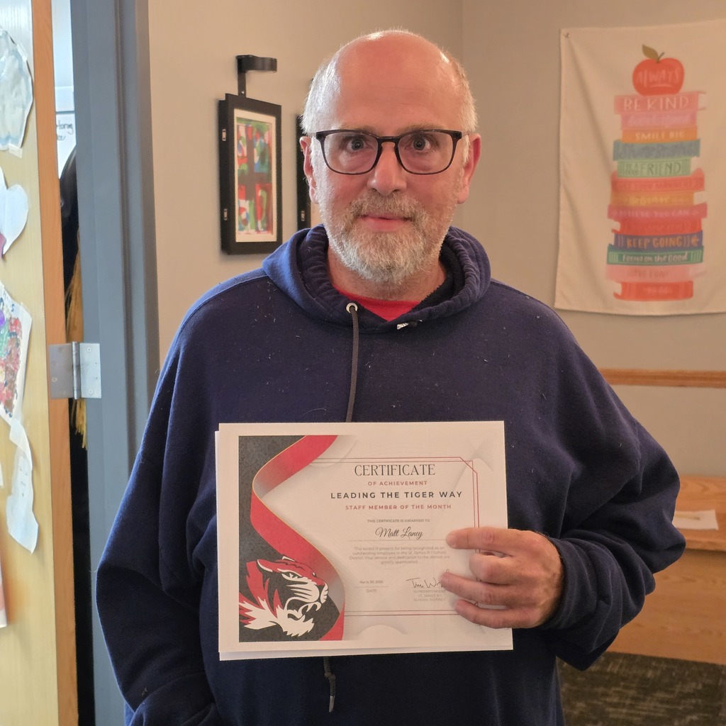 Man in a blue hoodie holds a certificate with a tiger graphic. Behind him is a wall with a banner and a framed picture.