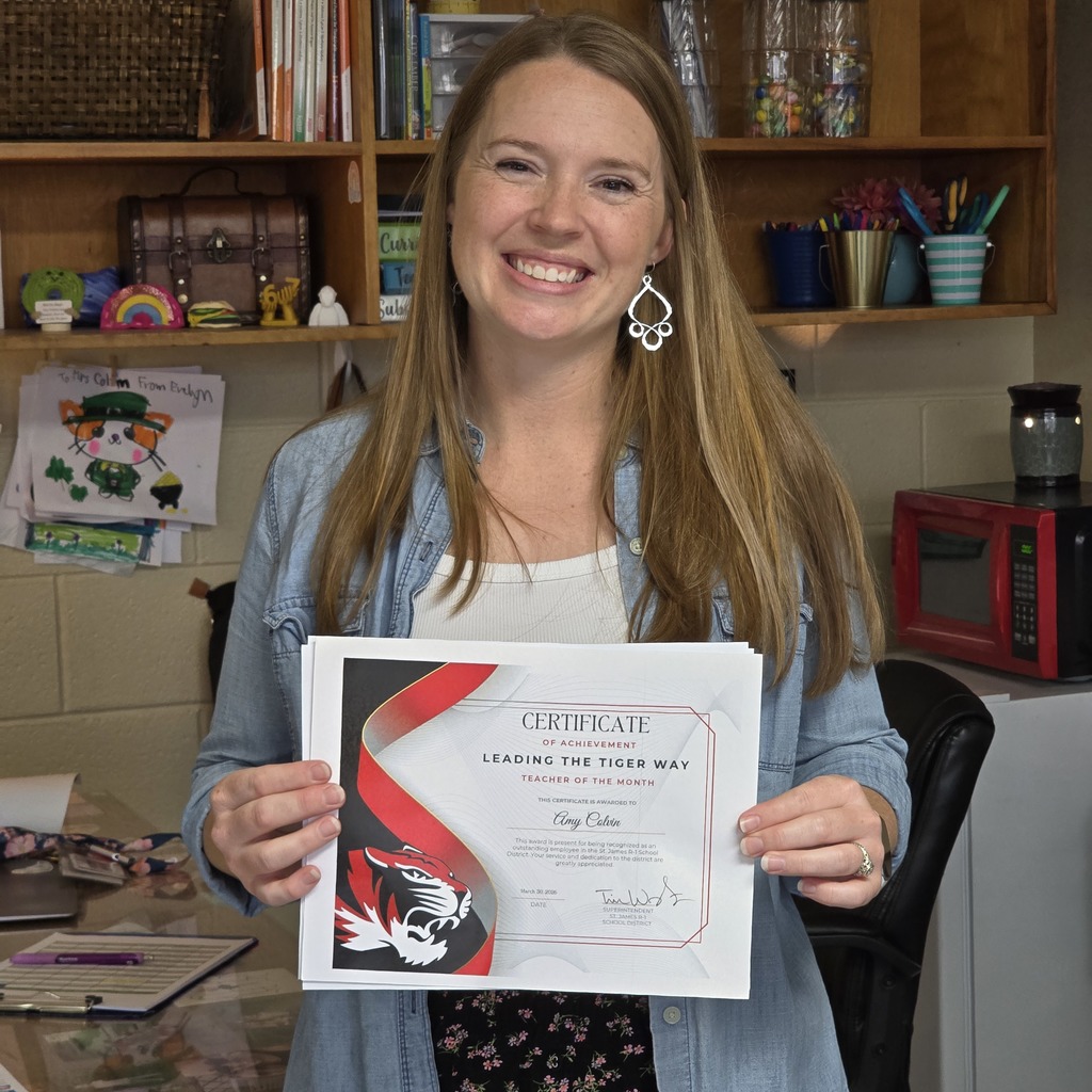 A woman holds a certificate in her hands while standing in an office. Behind her are shelves filled with items.