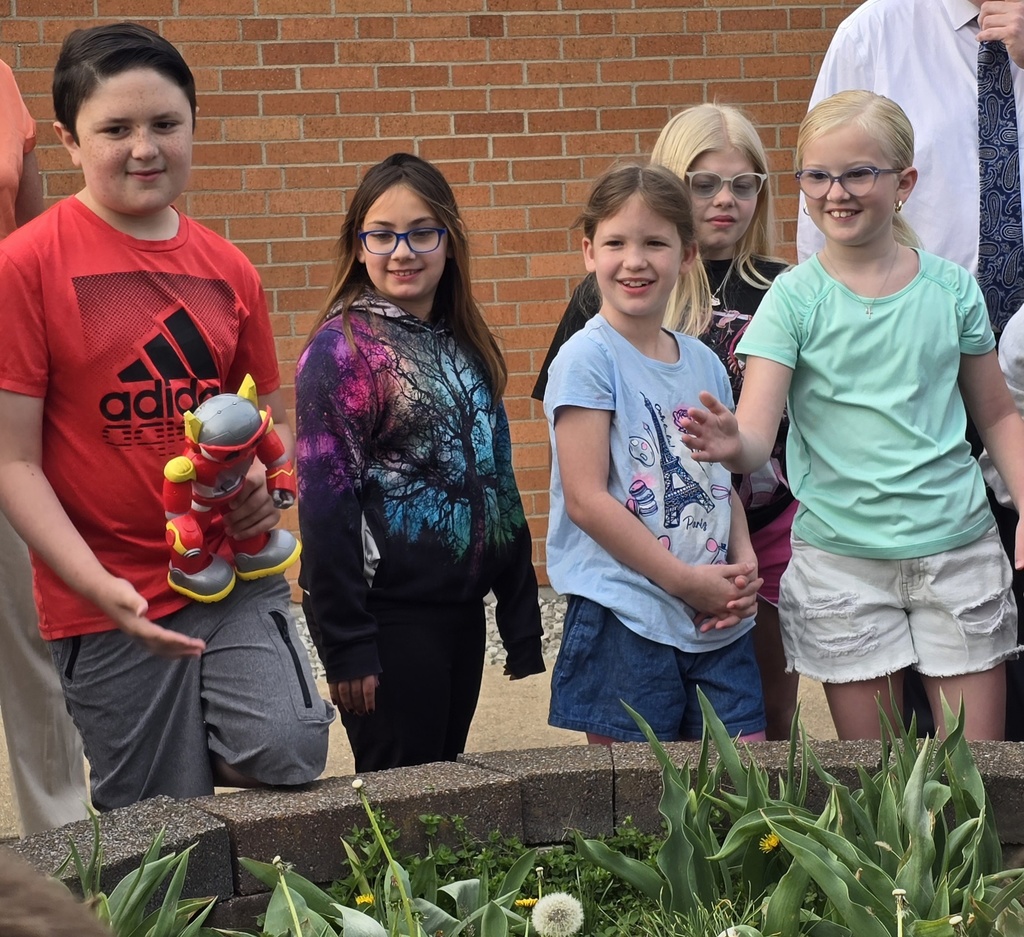 Five children stand together outside, with one holding a stuffed animal. Brick wall in background.